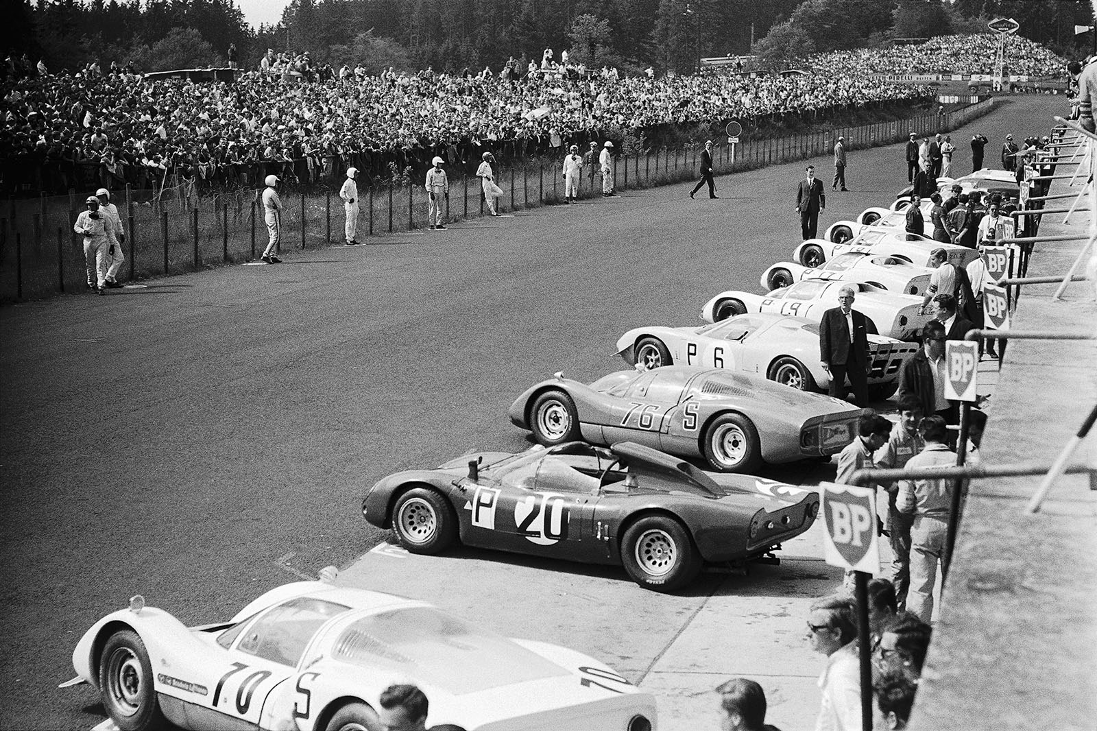 The race machines stand in line for the start of the 1000kms race whilst the drivers wait for the signal opposite their sports cars. From front: Scuderia Lufthansa's Porsche 906 with starting number 70, the Alfa Romeo T33 of Autodelta SpA (nr. 20), Sten Axelsson's (S) Porsche 906 #117 (nr. 76), the Mirage M1 Ford 10001 entered by J. W. Automotive Engineering, and a row of Porsches...