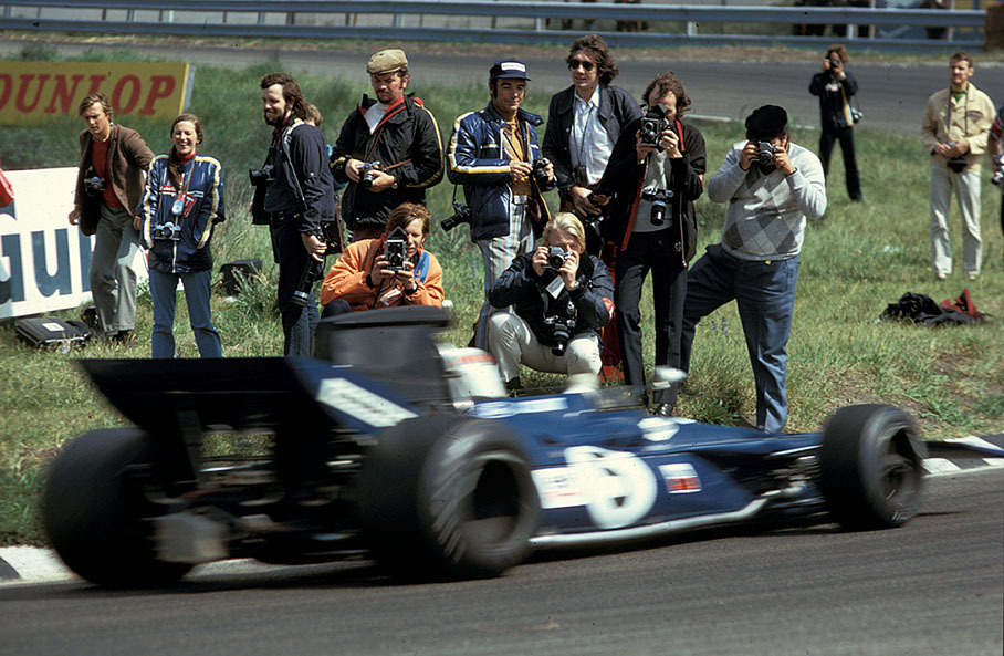 Photographers standing close to action, while Jackie Stewart (Tyrrell Ford-Cosworth) passes by.